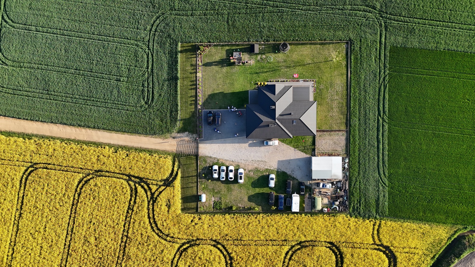 Zdjęcie z drona: nowoczesny dom z ciemnym dachem otoczony zielonym i żółtym polem, liczne samochody na podjeździe, symetryczny układ kompozycyjny.
