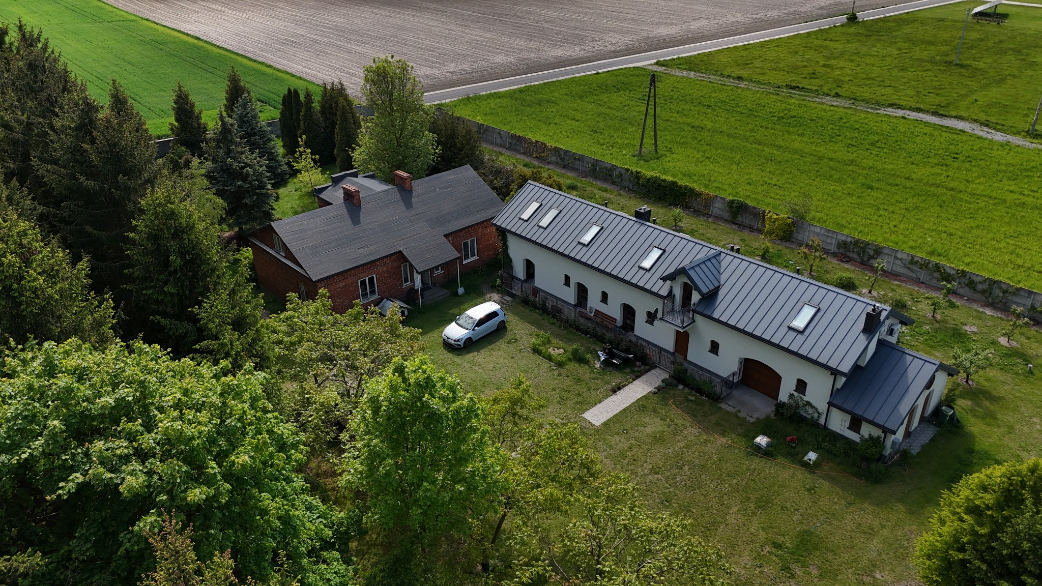 Zdjęcie z lotu ptaka przedstawia dwa budynki: ceglany dom z ciemnym dachem i biały budynek z szarym dachem, otoczone zielonym trawnikiem, drzewami i polami.