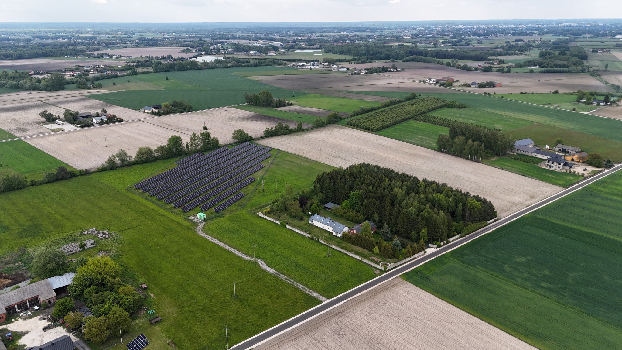 Szeroki, panoramiczny widok z lotu ptaka na wiejski krajobraz z polami, lasami, zabudowaniami i rząd paneli słonecznych w słoneczny dzień.