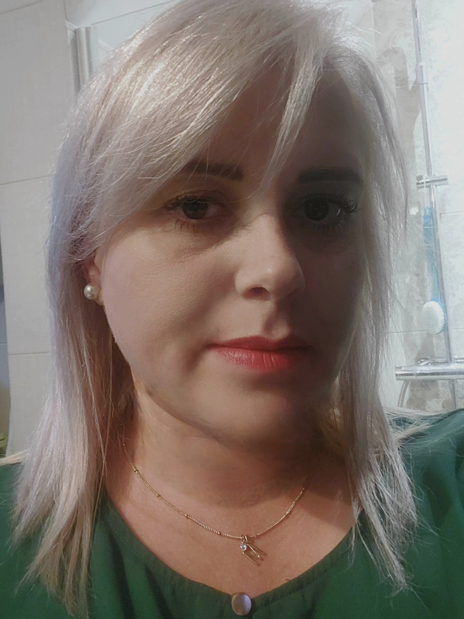 Portret kobiety z platynowymi włosami, w zielonej bluzce, z delikatnym makijażem i złotym łańcuszkiem, na tle łazienki z prysznicem.