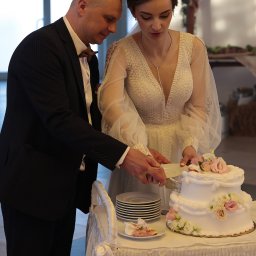 FotoMioduch - Para młoda kroi tort weselny, on w czarnym garniturze z muszką, ona w białej sukni z koronką i długimi rękawami, kwiaty na torcie.