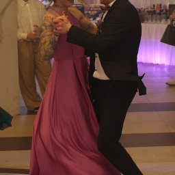 FotoMioduch - Uśmiechnięta para tańczy na parkiecie, kobieta w różowej sukni, mężczyzna w garniturze z muszką, w tle bufet i goście, dynamiczne ujęcie z wesela.