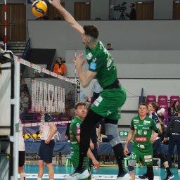 FotoMioduch - Dynamiczne ujęcie siatkarza w zielonym stroju podczas skoku do ataku na tle siatki i trybun. Widoczny numer '41' na koszulce, piłka w locie.