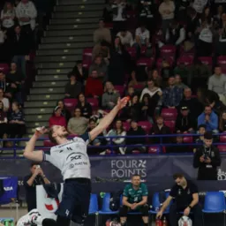 Dynamiczne ujęcie siatkarza w akcji, skaczącego do ataku na tle trybun wypełnionych kibicami. Widoczna piłka w locie i detale stroju sportowego.