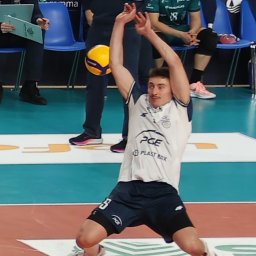 FotoMioduch - Mężczyzna w stroju sportowym odbija piłkę do siatkówki podczas meczu. W tle siedzący obserwatorzy i reklamy sponsorów.