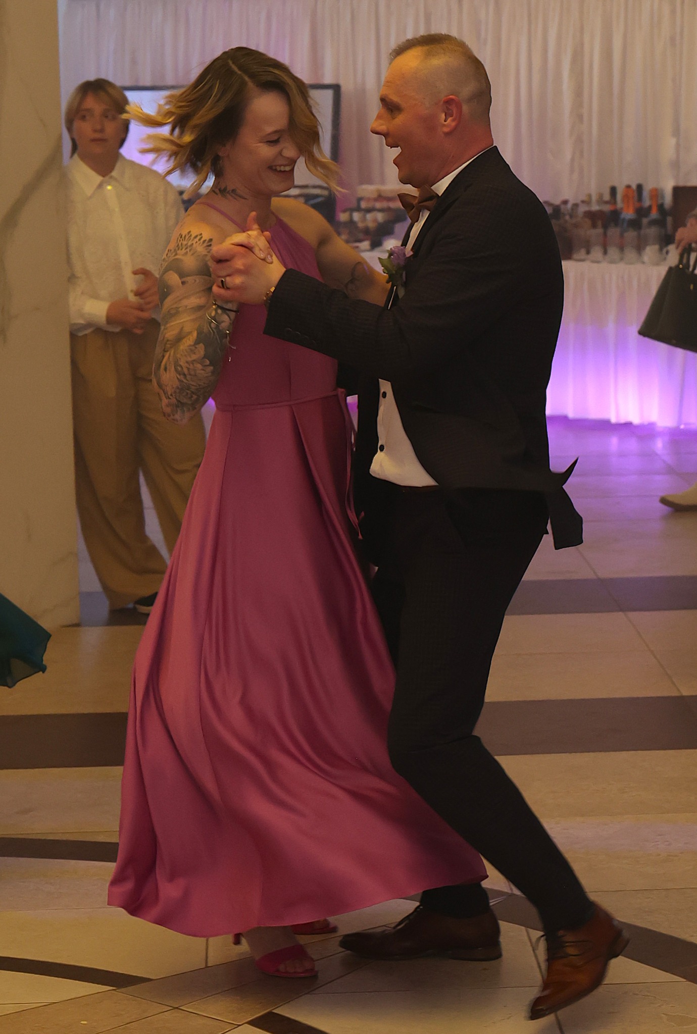 Uśmiechnięta para tańczy na parkiecie, kobieta w różowej sukni, mężczyzna w garniturze z muszką, w tle bufet i goście, dynamiczne ujęcie z wesela.