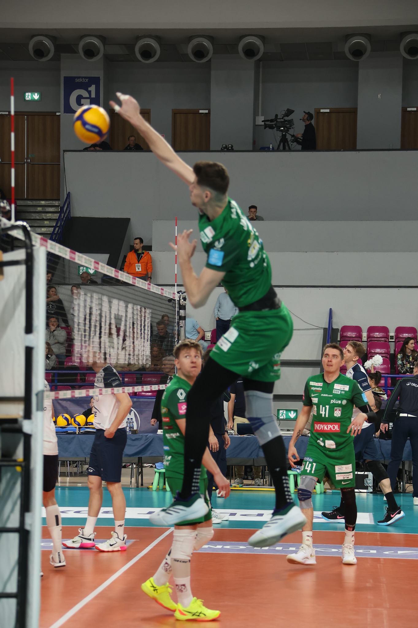 Dynamiczne ujęcie siatkarza w zielonym stroju podczas skoku do ataku na tle siatki i trybun. Widoczny numer '41' na koszulce, piłka w locie.