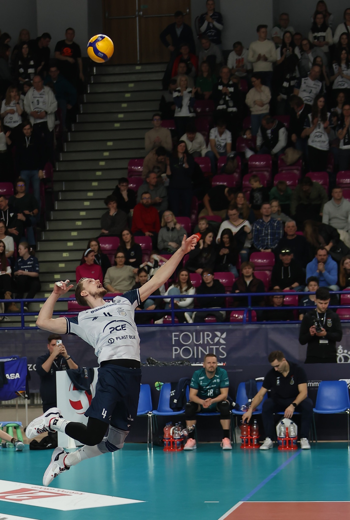 Dynamiczne ujęcie siatkarza w akcji, skaczącego do ataku na tle trybun wypełnionych kibicami. Widoczna piłka w locie i detale stroju sportowego.