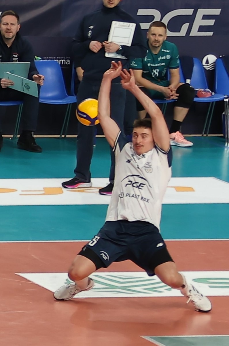 Mężczyzna w stroju sportowym odbija piłkę do siatkówki podczas meczu. W tle siedzący obserwatorzy i reklamy sponsorów.