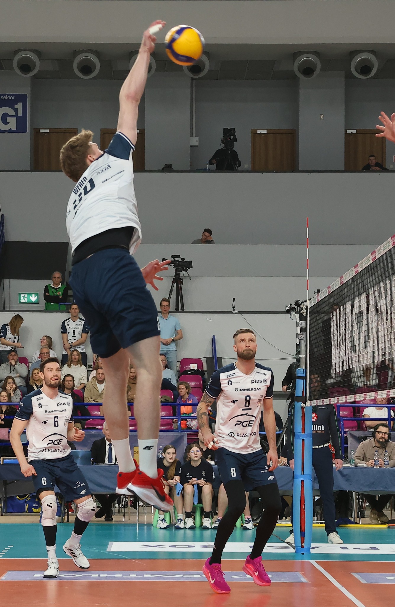 Dynamiczne ujęcie siatkarza w akcji, wyskakującego do ataku na tle trybun pełnych widzów podczas meczu. Widoczna siatka i oświetlenie hali sportowej.