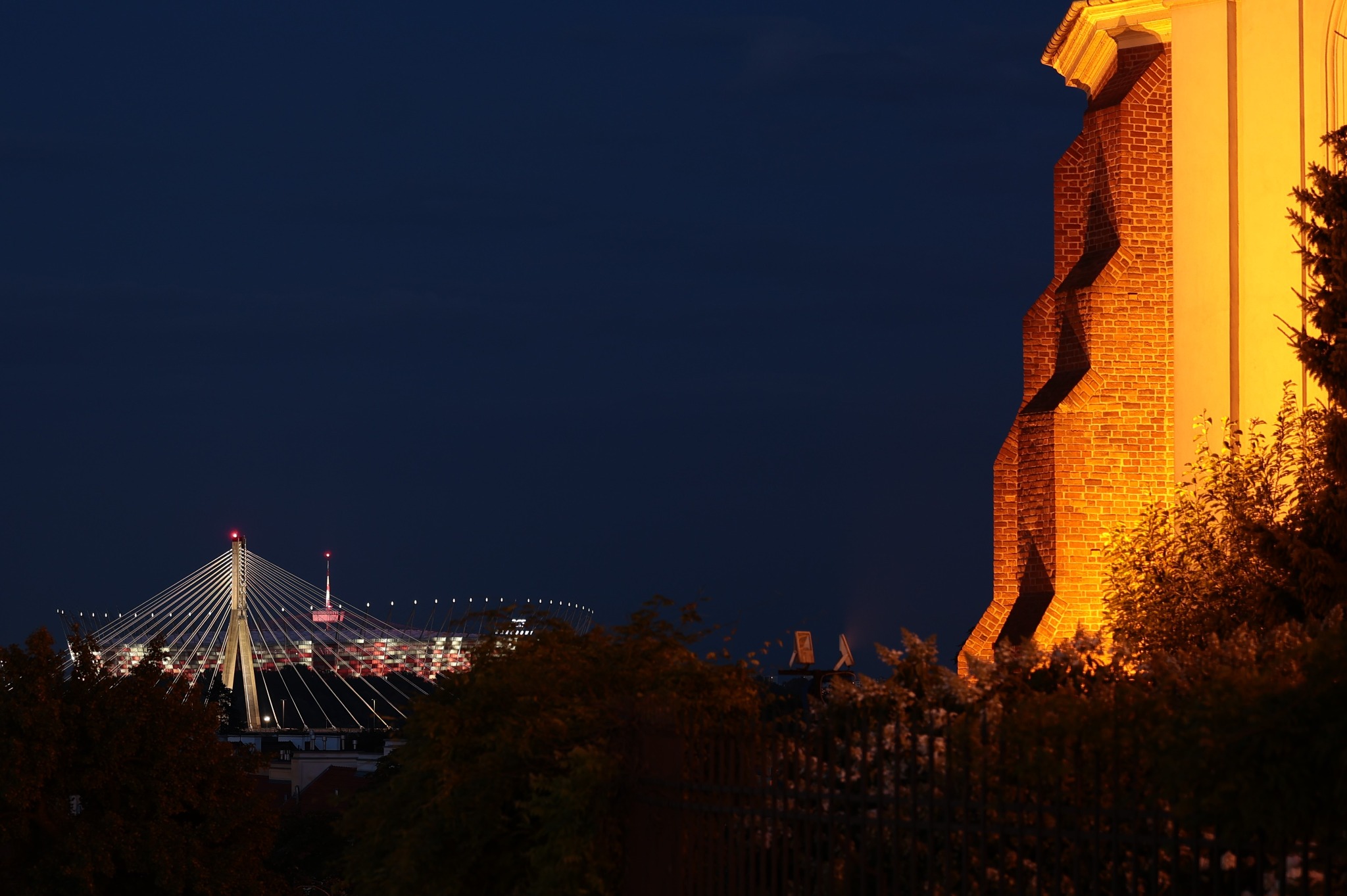 Nocny krajobraz miejski: oświetlony most linowy w oddali, ceglana wieża po prawej, ciemne niebo i zieleń w tle. Nietypowe ujęcie z dołu.