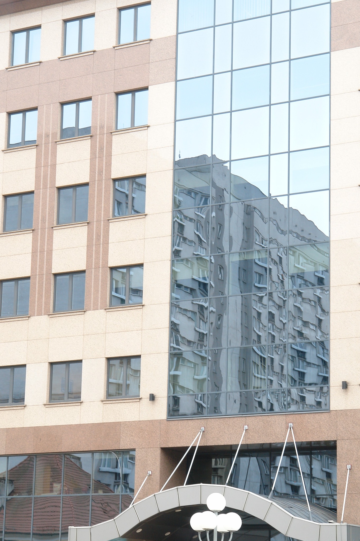 Budynek biurowy z fasadą z paneli w kolorze beżowym i dużymi oknami. W szklanej fasadzie odbija się sąsiedni budynek, tworząc iluzję optyczną.