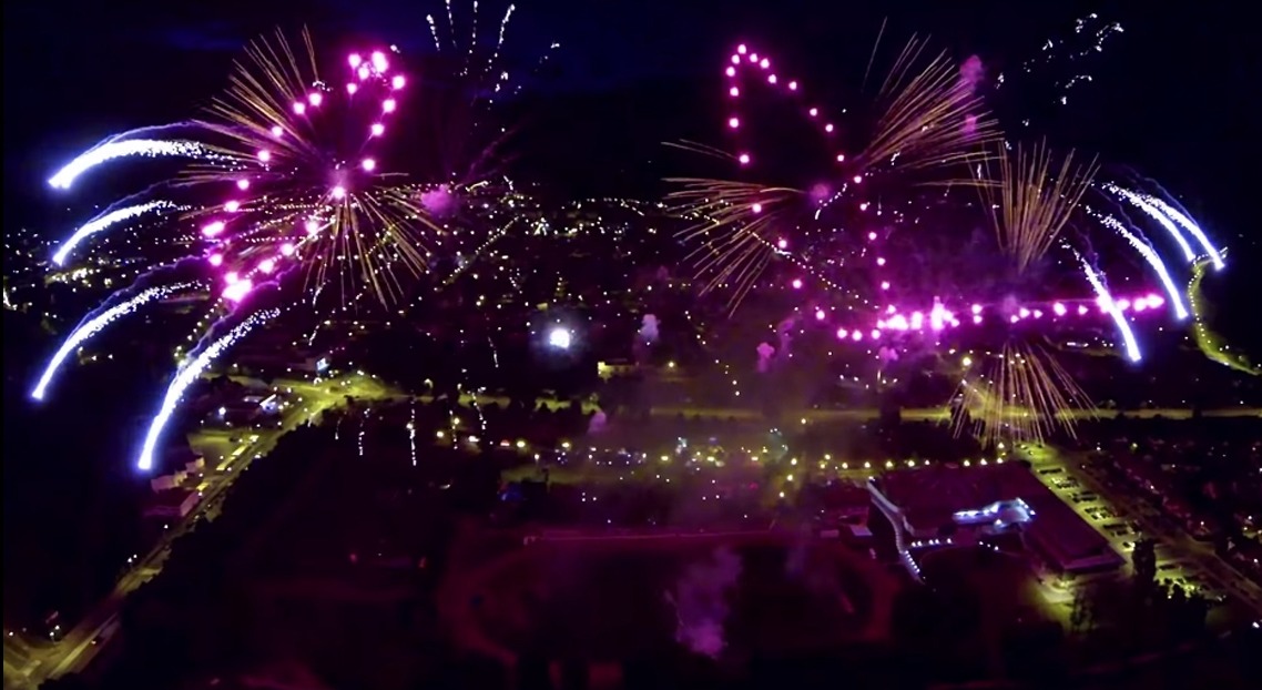 Nocne niebo rozświetlają symetryczne, różowo-złote fajerwerki nad miastem, widziane z lotu ptaka. Stadion i ulice tworzą tło dla pirotechnicznego pokazu.