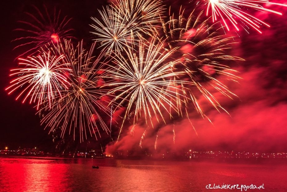 Spektakularny pokaz sztucznych ogni nad wodą, z czerwonymi refleksami na powierzchni. W tle widoczne światła miasta, a na pierwszym planie mała łódka.