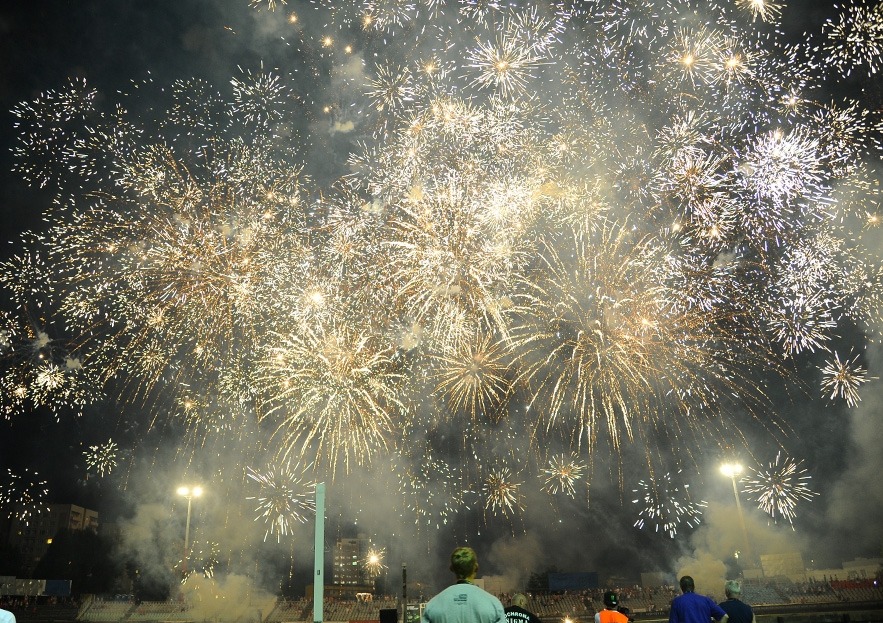Nocne niebo rozświetlone złotymi fajerwerkami nad stadionem, widziane z perspektywy publiczności. W tle budynki i oświetlenie stadionowe.
