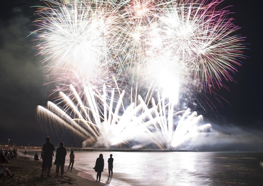 Nocny pokaz fajerwerków nad morzem, z sylwetkami ludzi na plaży. Jasne, białe i kolorowe eksplozje rozświetlają ciemne niebo nad wodą. Widoczny pomost w tle.