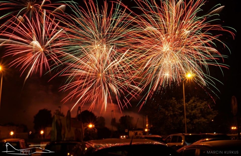 Nocne niebo rozświetlone feerią barw podczas pokazu pirotechnicznego, z widocznymi lampami ulicznymi i sylwetkami drzew w tle.