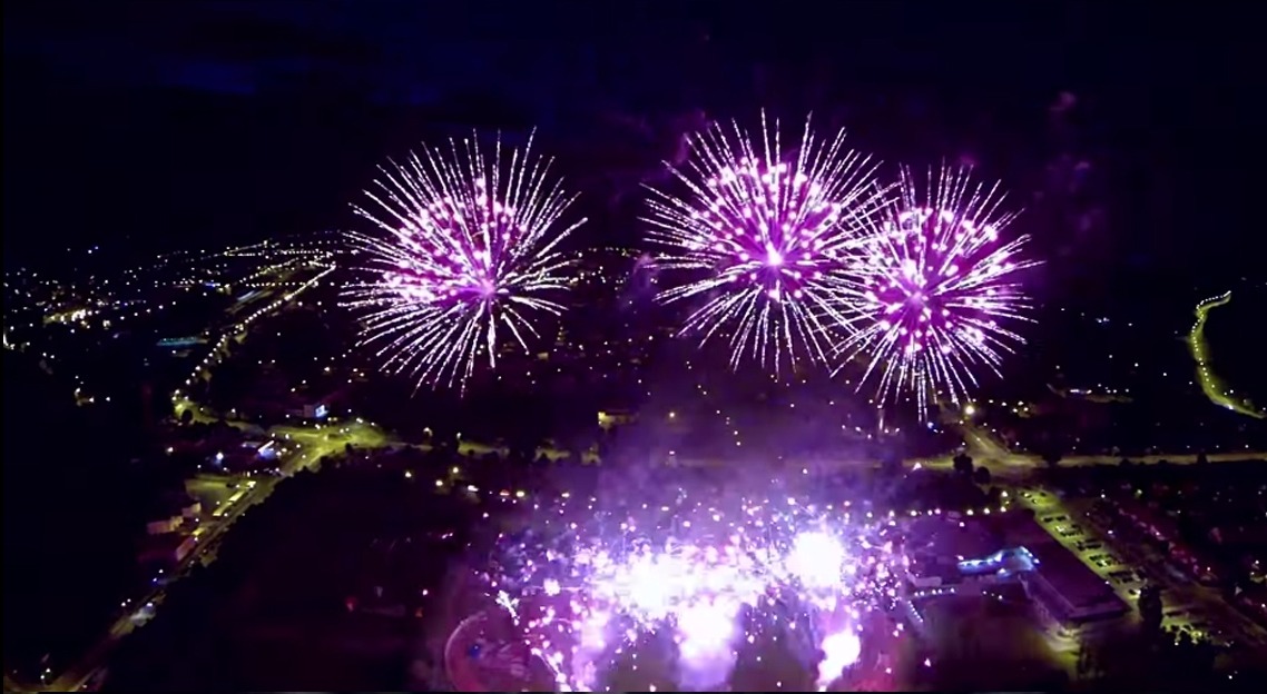 Nocny pokaz pirotechniczny nad miastem, fioletowe fajerwerki rozświetlają ciemne niebo nad zabudowaniami i drogami.