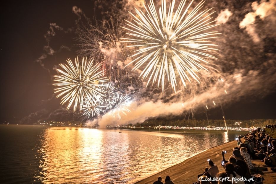 Nocny pokaz sztucznych ogni nad wodą, z odbiciami w tafli jeziora i sylwetkami widzów na brzegu. Jasne eksplozje rozświetlają niebo, tworząc spektakularne widowisko.