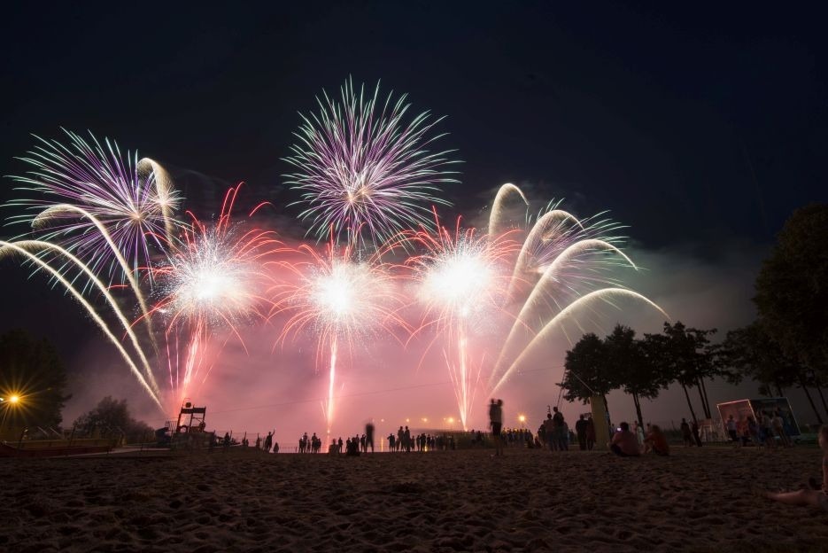 Spektakularny pokaz fajerwerków nad plażą w nocnej scenerii, z widocznymi sylwetkami obserwujących osób i drzewami w tle.
