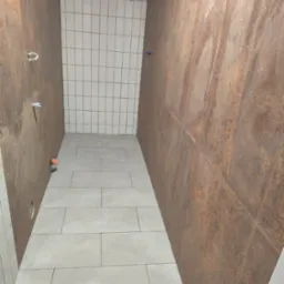 Wykończona łazienka z beżowymi płytkami imitującymi rdzę na ścianach i jasnymi, prostokątnymi płytkami na podłodze i części ściany pod sufitem. Widoczne instalacje.