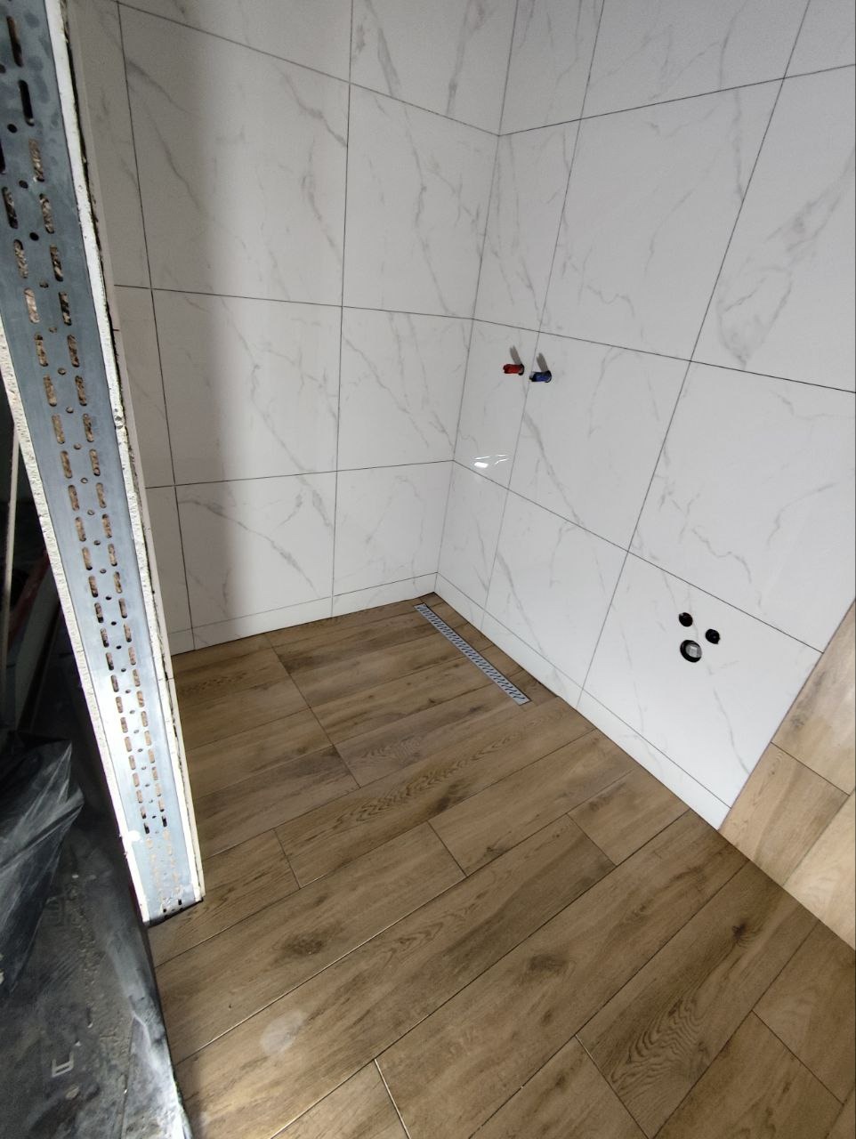 Wykończona łazienka z marmurowymi płytkami na ścianach i drewnopodobną podłogą. Widoczny odpływ liniowy i wystające rury instalacji wodnej. Część regału w lewym rogu.