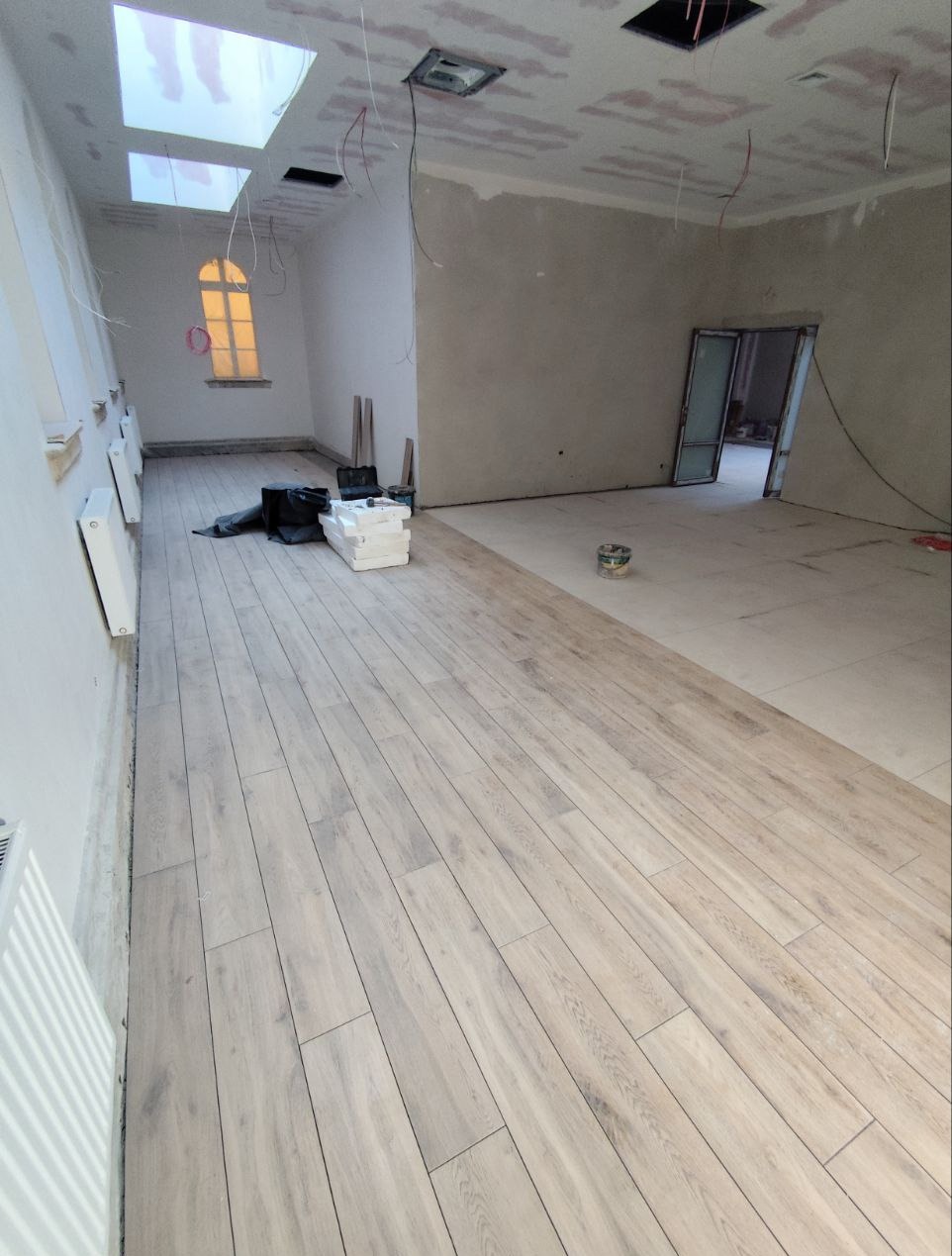 Parkiet ułożony w dużym pokoju z oknem łukowym i surowymi ścianami. Część podłogi wyłożona płytkami. Widoczne kable i materiały budowlane.