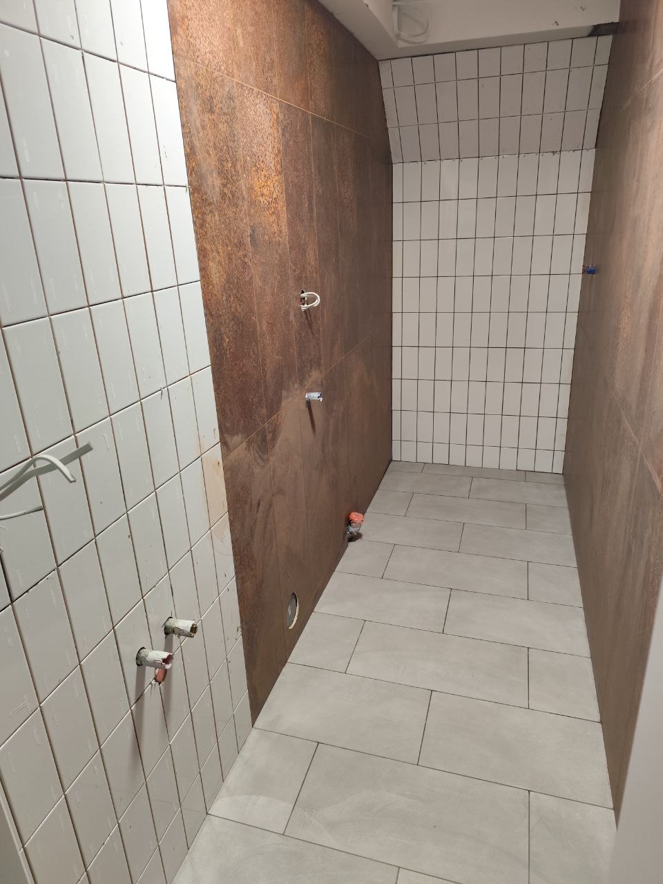 Wnętrze łazienki w trakcie remontu: białe i rdzawobrązowe płytki na ścianach, szare płytki podłogowe, wystające rury. Widoczne etapy prac wykończeniowych.