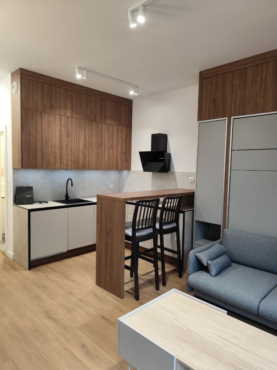 Nowoczesna kuchnia z drewnianymi szafkami, szarymi płytkami heksagonalnymi i blatem barowym z krzesłami. Minimalistyczny design, oświetlenie punktowe.