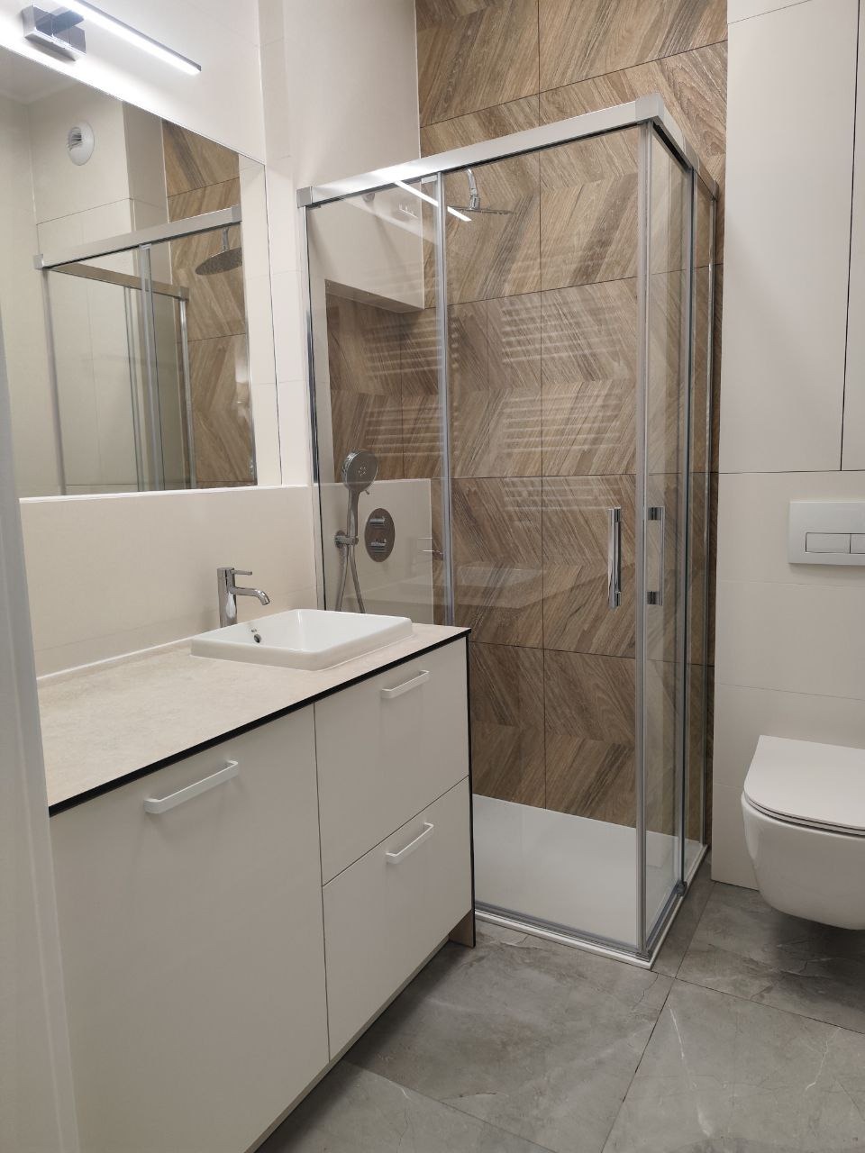 Nowoczesna łazienka z kabiną prysznicową, umywalką w zabudowie i toaletą. Brązowe płytki z wzorem jodełki na ścianie kabiny. Szare płytki podłogowe.