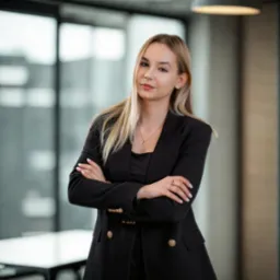 Profesjonalny portret kobiety w czarnej marynarce na tle biura. Skrzyżowane ręce, subtelny uśmiech, elegancka biżuteria. Jasne włosy, naturalny makijaż, nowoczesne wnętrze.