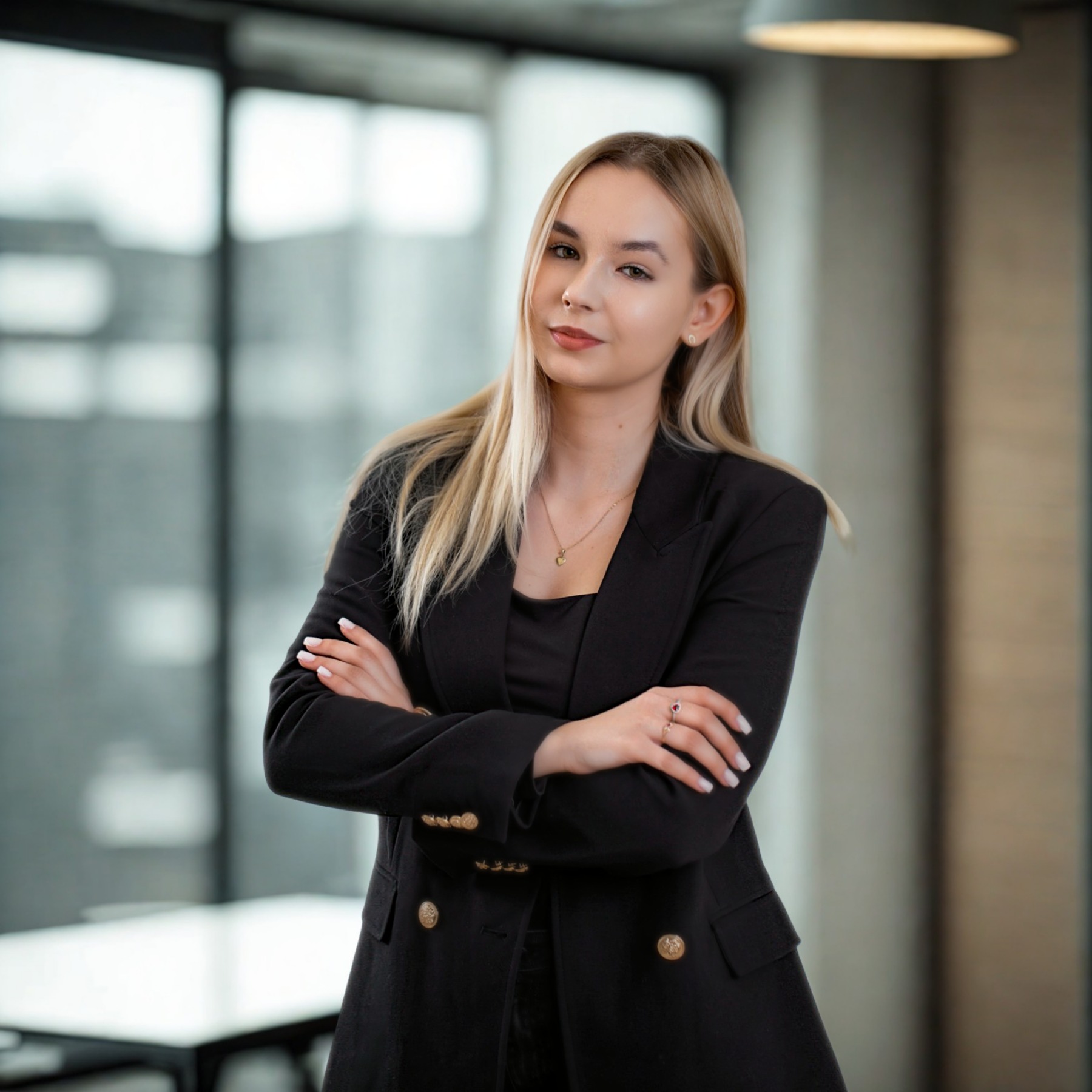 Profesjonalny portret kobiety w czarnej marynarce na tle biura. Skrzyżowane ręce, subtelny uśmiech, elegancka biżuteria. Jasne włosy, naturalny makijaż, nowoczesne wnętrze.