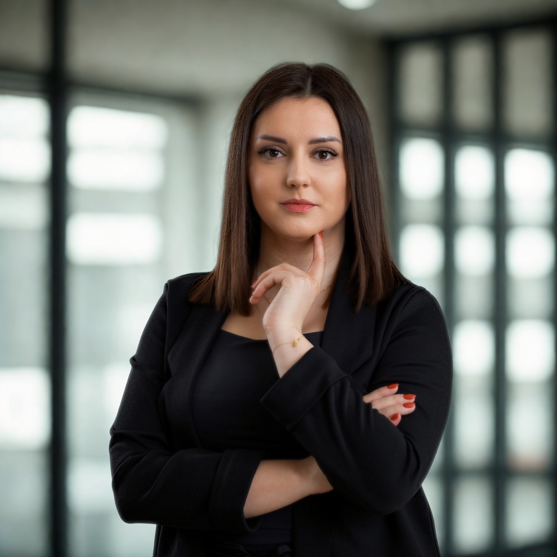 Profesjonalny portret kobiety w czarnym żakiecie, z ręką pod brodą, wyrażający pewność siebie. W tle biurowe okna z rozmytym światłem.