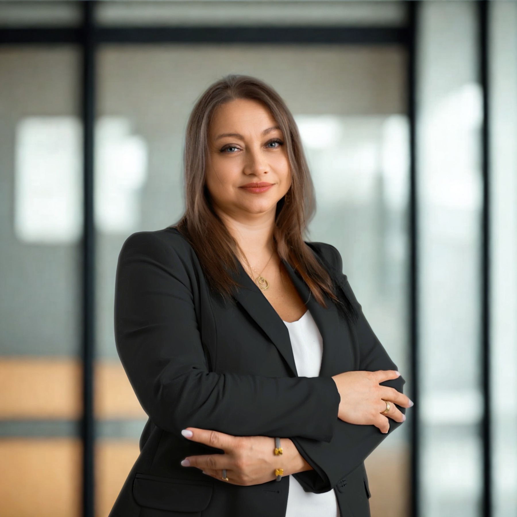 Portret kobiety w czarnej marynarce, z założonymi rękami, na tle przeszklonej ściany biurowej. Profesjonalny wizerunek.