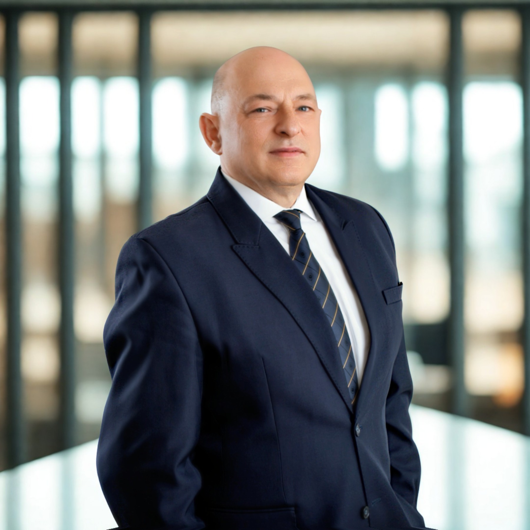 Portret biznesowy mężczyzny w granatowym garniturze i krawacie w paski, na rozmytym tle biurowym z oknami. Profesjonalny wizerunek.