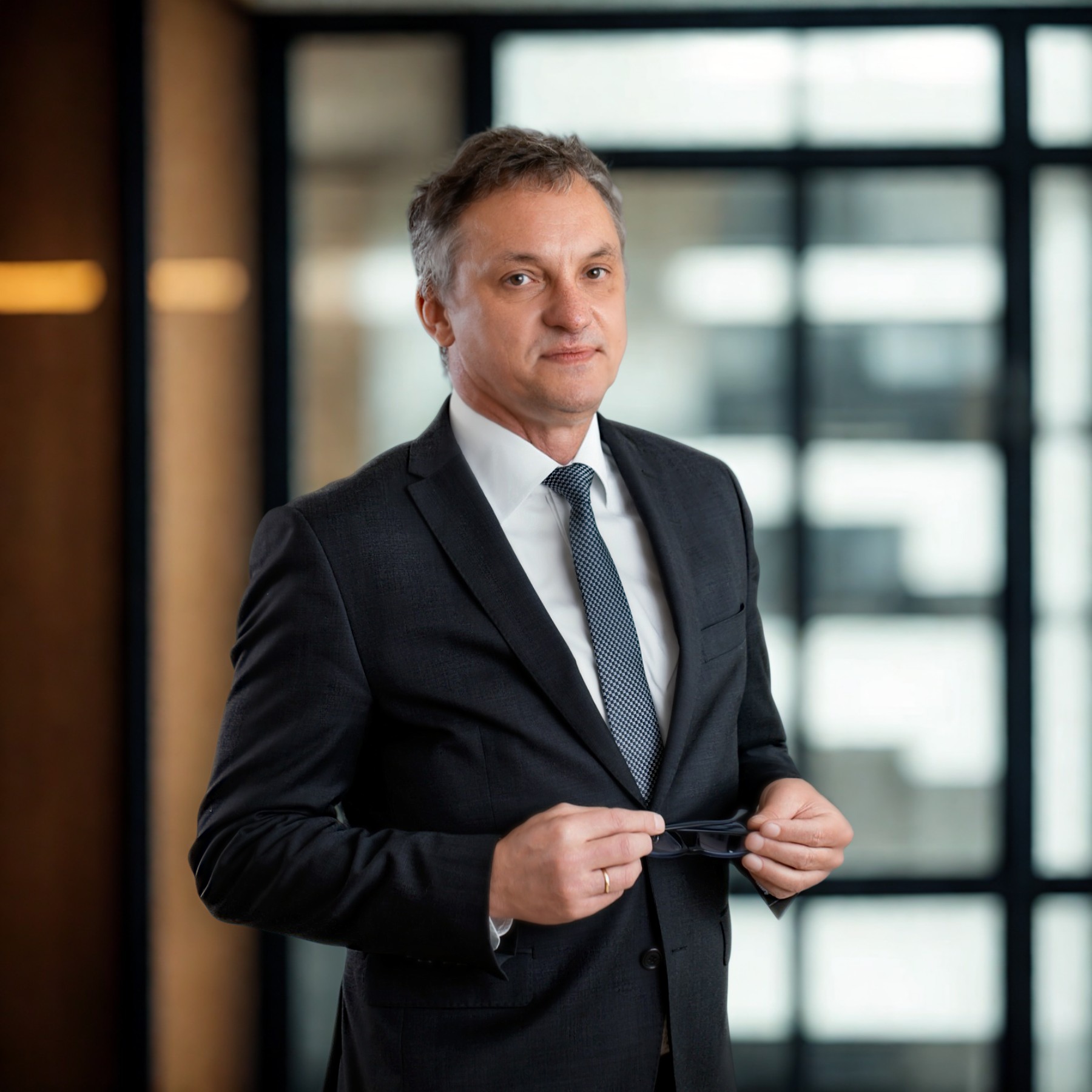 Portret mężczyzny w garniturze, trzymającego okulary na tle nowoczesnego biura z przeszkleniami i drewnianymi elementami. Profesjonalny wygląd.