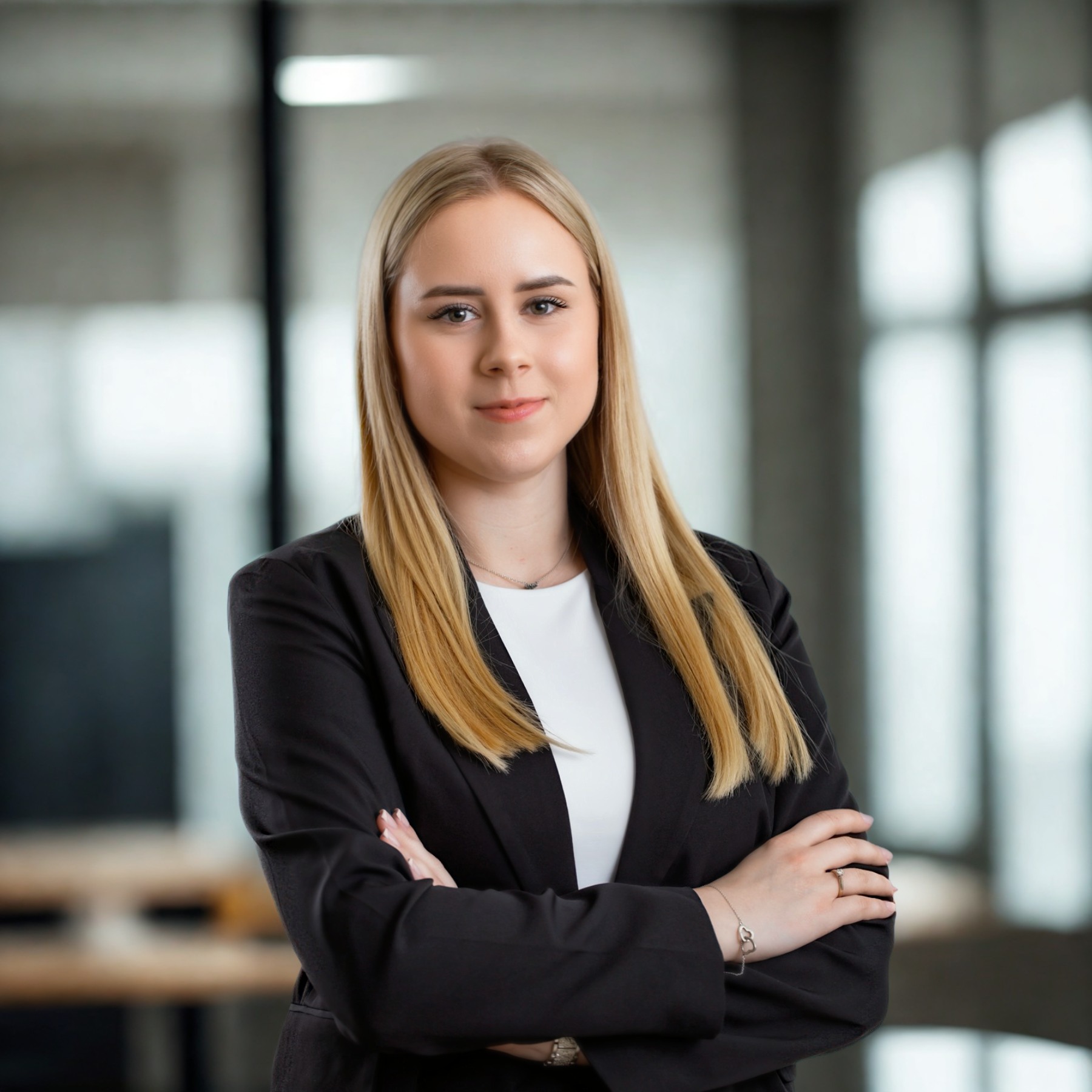 Portret młodej kobiety w czarnej marynarce, z blond włosami i skrzyżowanymi rękami, na rozmytym tle biurowym z oknem i szklaną ścianą.
