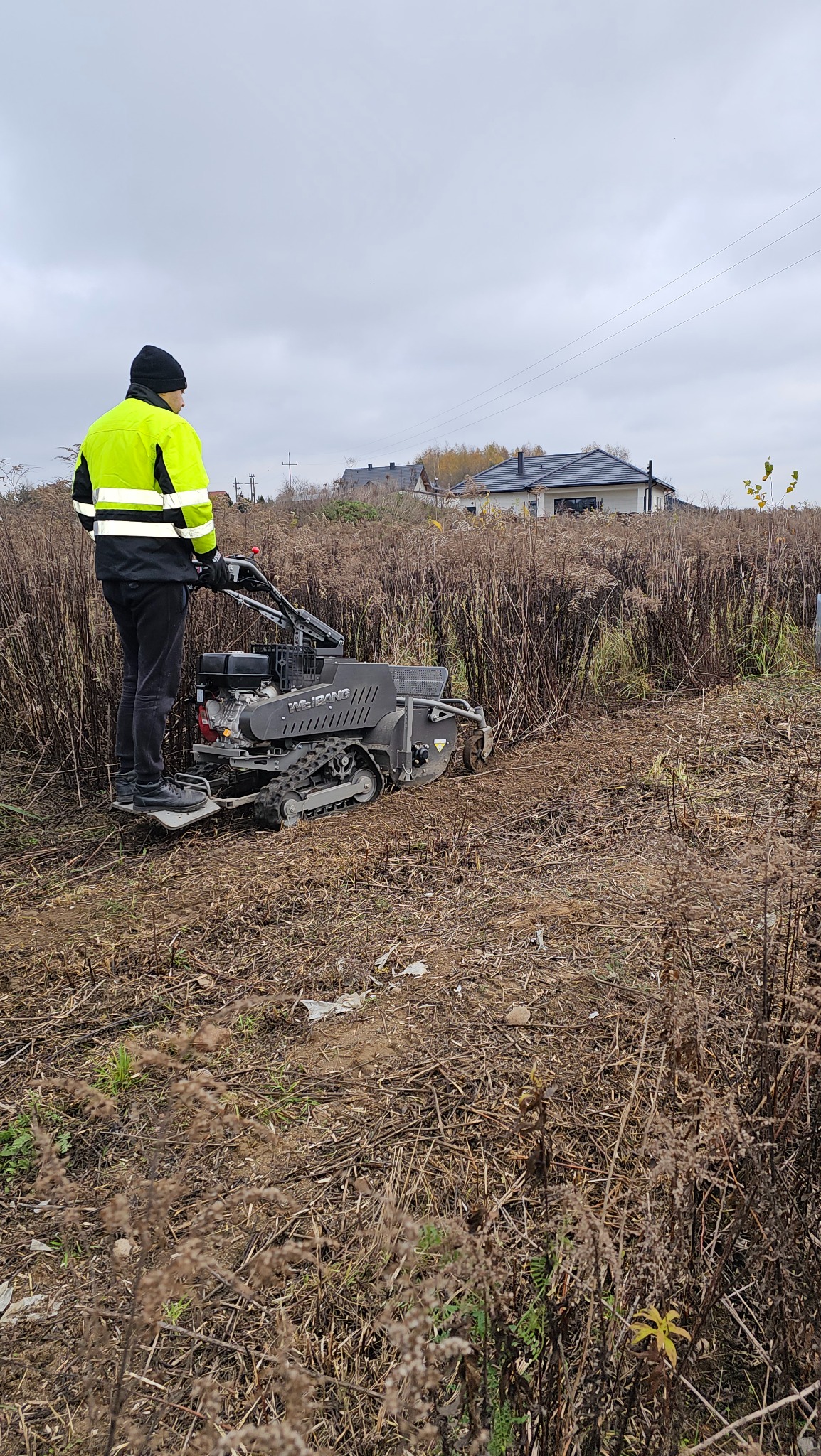 Operator w odblaskowej kurtce obsługuje glebogryzarkę na gąsienicach, przygotowując teren pod uprawę. W tle zabudowa jednorodzinna i pochmurne niebo.