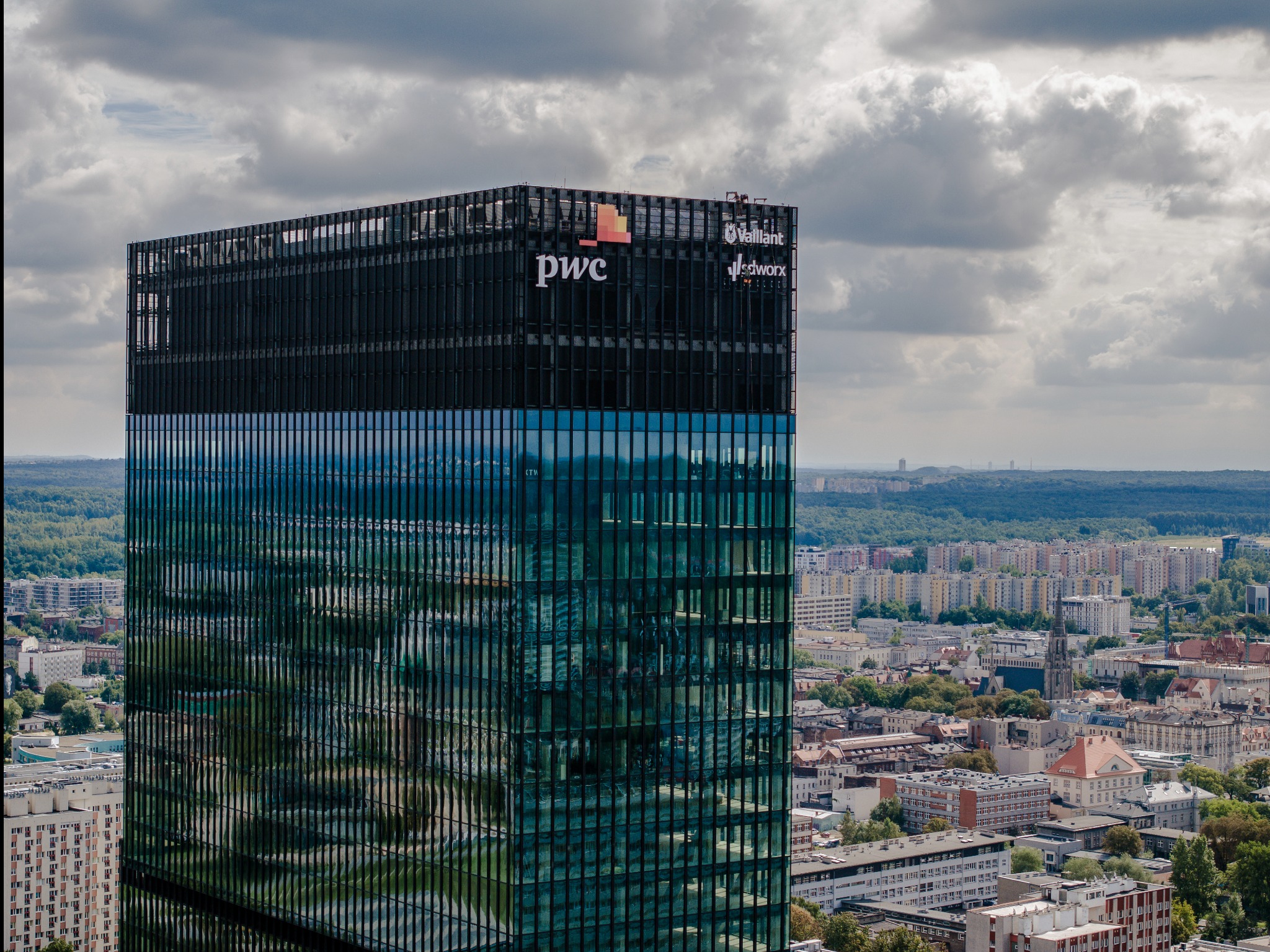 Wysoki biurowiec z logo PWC i Vaillant na tle miejskiego krajobrazu pod pochmurnym niebem. Widok z góry na miasto z budynkami i zielenią.