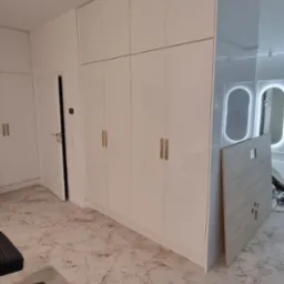 Biała szafa wnękowa z złotymi uchwytami, dopasowana do sufitu, na tle marmurowej podłogi. Widoczne lustra z podświetleniem i fragmenty mebli.