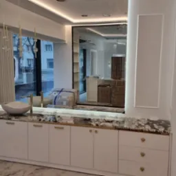 Biała szafka łazienkowa z marmurowym blatem i złotymi uchwytami, okrągła umywalka, lustro z oświetleniem, widok na ulicę przez okno. Elegancki design.