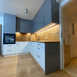 P&K Meble - Nowoczesna kuchnia z szarymi szafkami górnymi i białymi dolnymi, drewnianym blatem i oświetleniem LED. Widoczny piekarnik i czarna armatura.