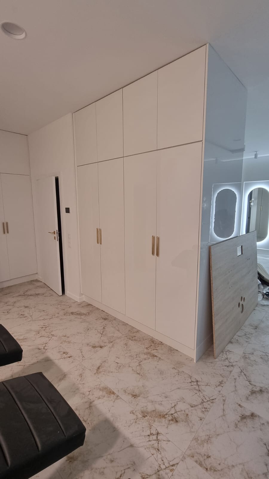 Biała szafa wnękowa z złotymi uchwytami, dopasowana do sufitu, na tle marmurowej podłogi. Widoczne lustra z podświetleniem i fragmenty mebli.