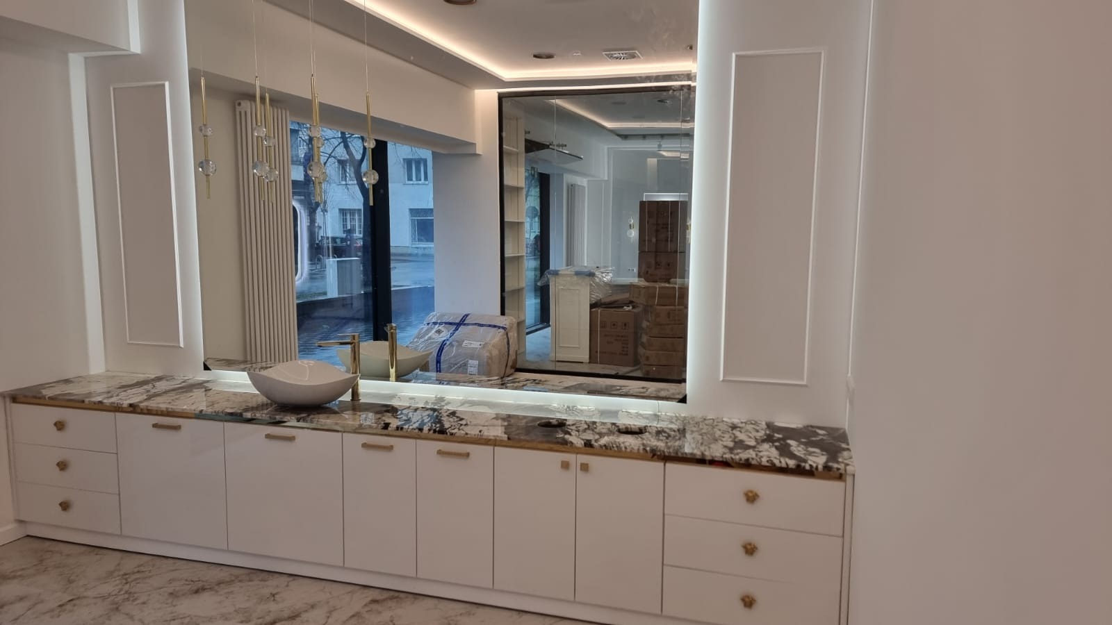 Biała szafka łazienkowa z marmurowym blatem i złotymi uchwytami, okrągła umywalka, lustro z oświetleniem, widok na ulicę przez okno. Elegancki design.