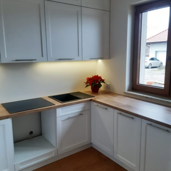 Jasna kuchnia na wymiar z drewnianym blatem, czarnym zlewem i płytą. Na blacie czerwona gwiazda betlejemska. Widok na dom i auto przez okno.