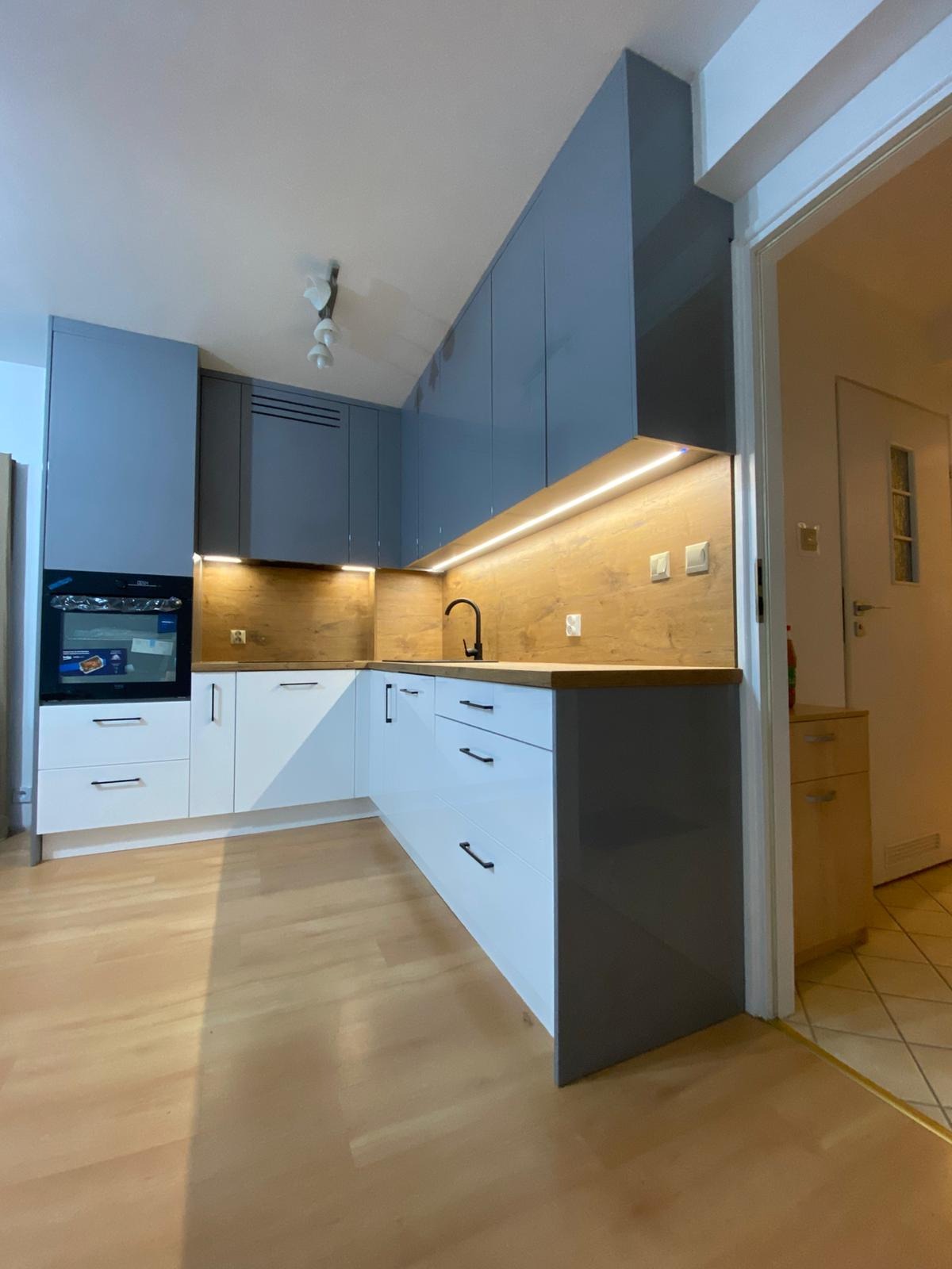 Nowoczesna kuchnia z szarymi szafkami górnymi i białymi dolnymi, drewnianym blatem i oświetleniem LED. Widoczny piekarnik i czarna armatura.