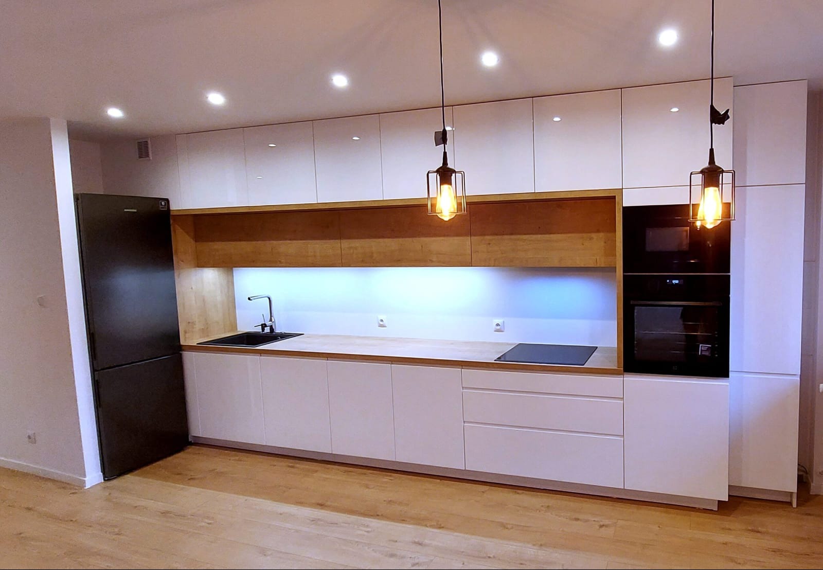 Nowoczesna kuchnia z białymi frontami, drewnianymi akcentami i czarnym sprzętem AGD. Wiszące lampy z żarówkami Edisona dopełniają aranżację.