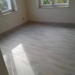 OFERTEO S.A - Jasne panele podłogowe w pustym pokoju z dwoma oknami, widok na drzewa. Świeżo wykończone wnętrze po remoncie, gotowe do umeblowania.