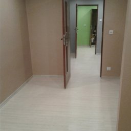 OFERTEO S.A - Jasne panele podłogowe w pustym pokoju z beżowymi ścianami. Otwarte drzwi ukazują dalszą część mieszkania z zielonymi ścianami.