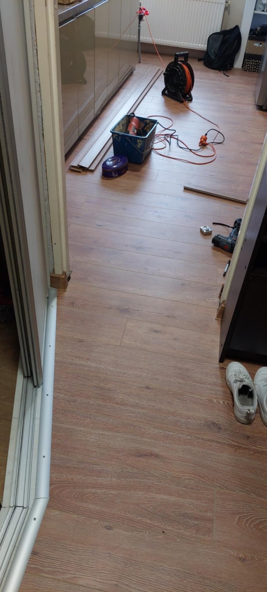 Podłoga po remoncie: panele ułożone, listwy przypodłogowe zamontowane. Widoczne narzędzia i akcesoria malarskie. Buty robocze w rogu.